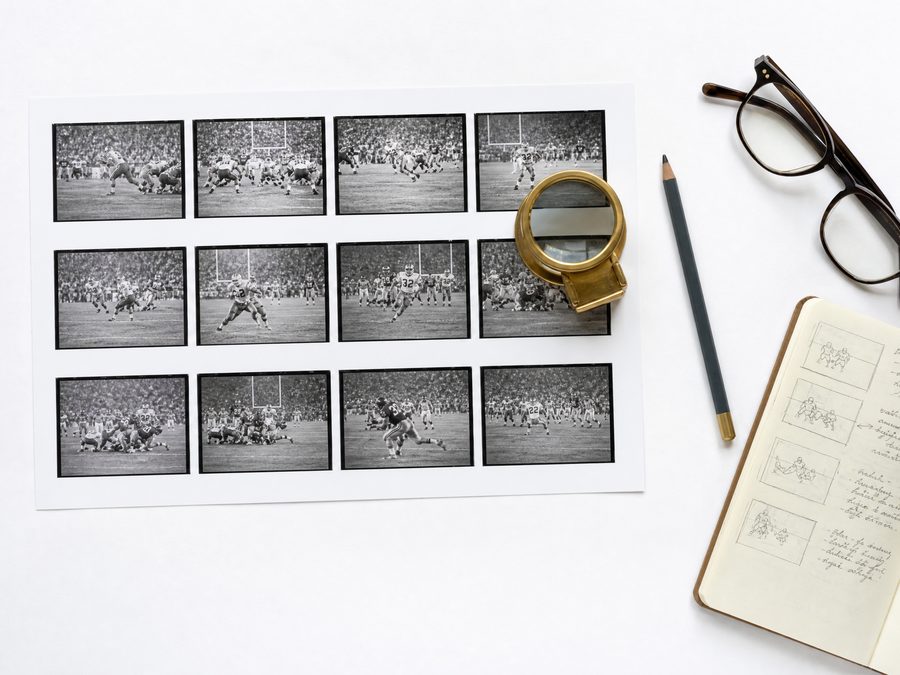 An overhead view of a printed contact sheet of small game frames on a white desk with a brass loupe resting on one frame, a grease pencil and a notebook of composition sketches
